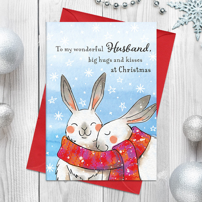 Wonderful Husband Christmas card featuring two illustrated rabbits snuggling together in a shared patterned scarf, surrounded by snowflakes and stars, with the greeting ‘Big hugs and kisses at Christmas
