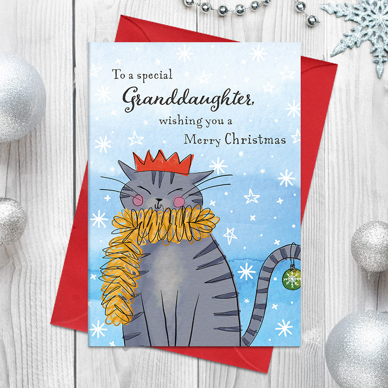 Special Granddaughter Christmas card featuring a smiling striped cat wearing a party crown and tinsel scarf, with a bauble hanging from its curled tail, surrounded by snowflakes and stars