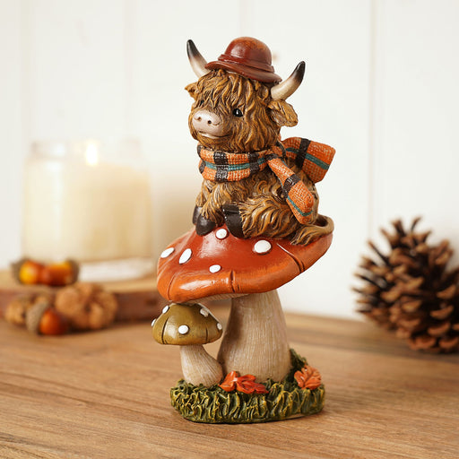 Highland cow figurine on rust-coloured spotted toadstool, wearing bowler hat and orange tartan scarf, shaggy fur and curved horns, autumn lifestyle shot on wooden surface