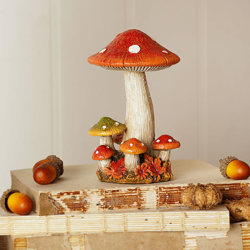 A resin mushroom cluster ornament with a tall central red-capped mushroom surrounded by smaller mushrooms, styled on a rustic wooden surface with acorns and autumn foliage against a soft neutral background
