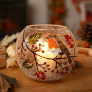 A round crackle glass votive holder with a hand-painted robin and autumn berry branch, glowing with a warm lit tealight, styled on a wooden surface with pinecones and autumn leaves in the background