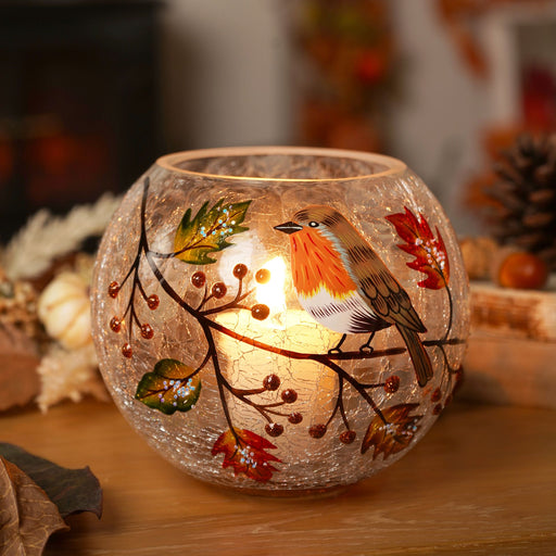 A round crackle glass votive holder with a hand-painted robin and autumn berry branch, glowing with a warm lit tealight, styled on a wooden surface with pinecones and autumn leaves in the background