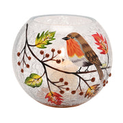 A round crackle glass votive holder with a hand-painted robin perched on a berry branch surrounded by autumn leaves, glowing warmly with a lit tealight inside, front view on a white background