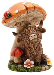 Highland Cow Toadstool Figurine, shaggy highland cow sitting beside large spotted toadstool with ladybird on cap, small red toadstool and wildflowers at base, front view on white background