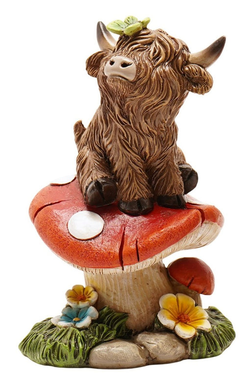 Highland Cow Sitting on Toadstool Figurine, shaggy highland cow perched on large spotted toadstool with butterfly on head, smaller toadstool and wildflowers at base, front view on white background