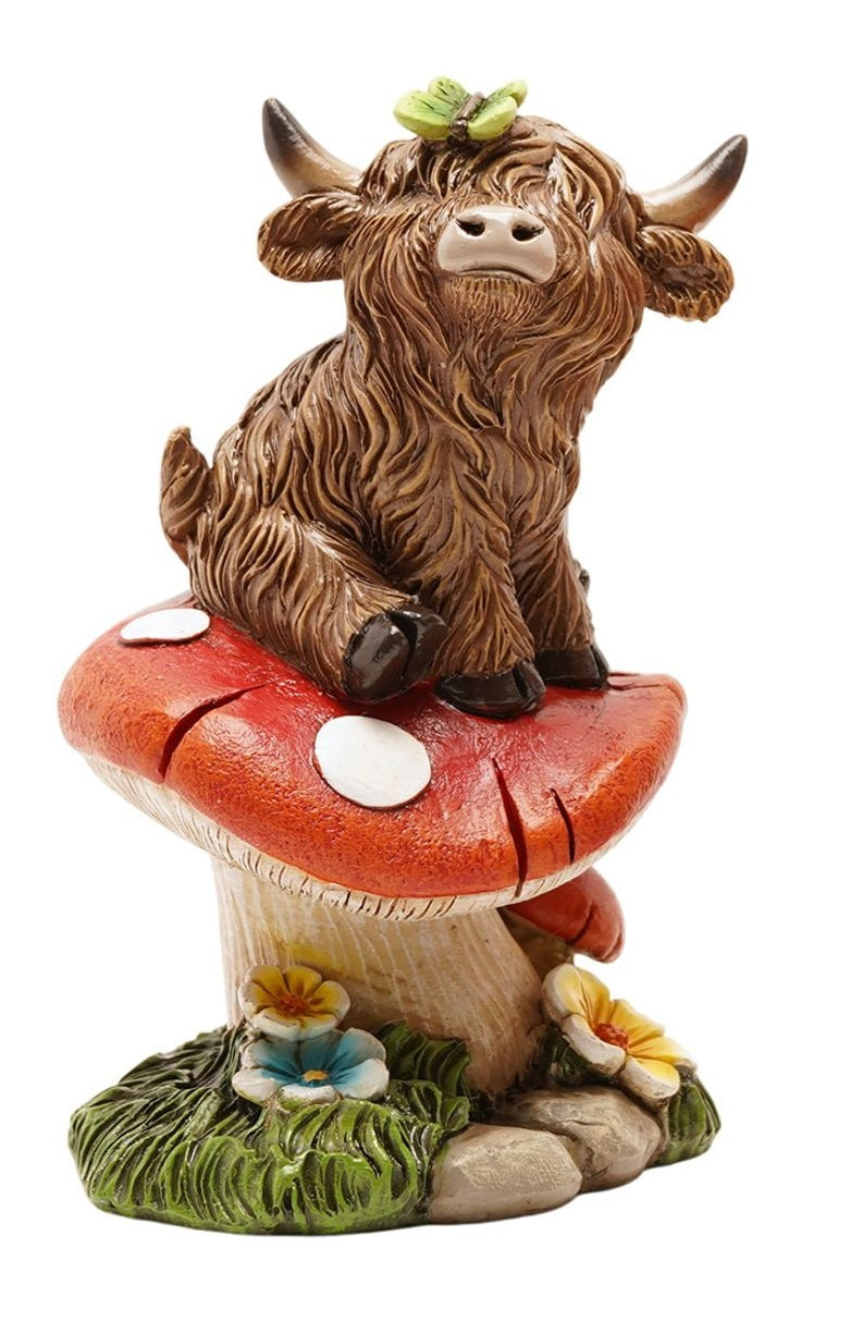 Highland Cow Sitting on Toadstool Figurine side view showing highland cow with curved horns and butterfly seated on spotted toadstool with wildflowers at base, on white background