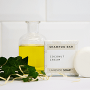 Laneside Soap Co Coconut Cream Shampoo Bar 60g with its box styled on a bathroom shelf with green foliage and a glass bottle of oil