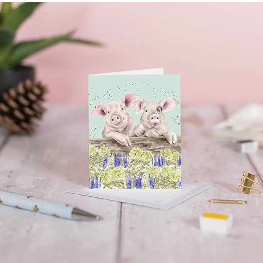 Wrendale Designs Nosy Neighboars pig card displayed upright on a wooden surface alongside a pine cone, pen and paper clips, with envelope