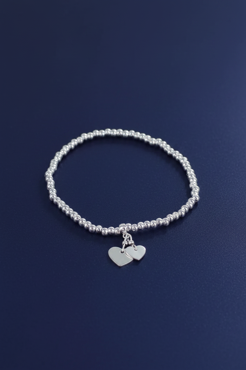 Silver plated elasticated bracelet with two dangling silver heart charms on a ball bead band, laid flat on a deep navy blue background
