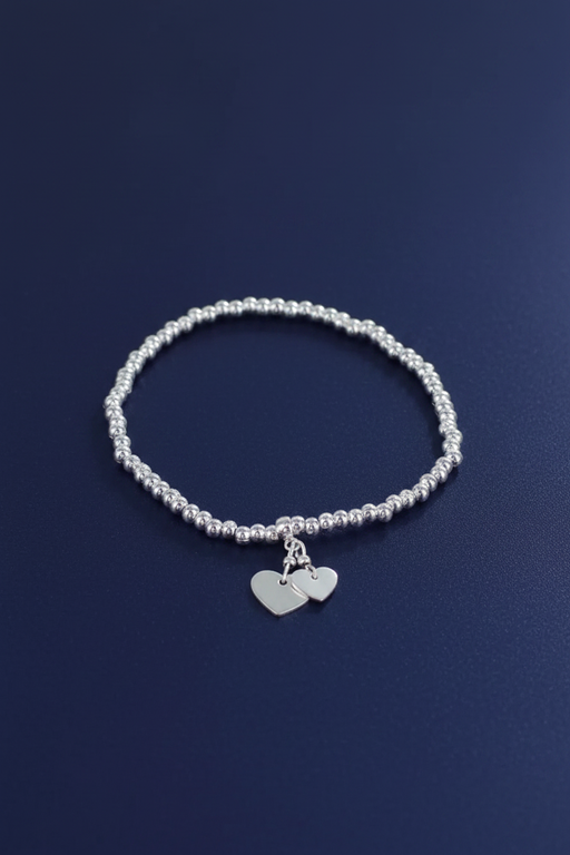Silver plated elasticated bracelet with two dangling silver heart charms on a ball bead band, laid flat on a deep navy blue background