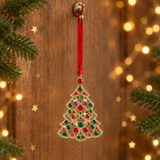 Gold plated crystal tree decoration hanging by a long red ribbon threaded through a rustic gold hook, surrounded by evergreen sprigs and gold star confetti with warm golden bokeh fairy lights in the background