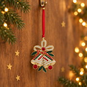 Gold plated crystal bells decoration hanging by a long red ribbon threaded through a rustic gold hook, surrounded by evergreen sprigs and gold star confetti with warm golden bokeh fairy lights in the background