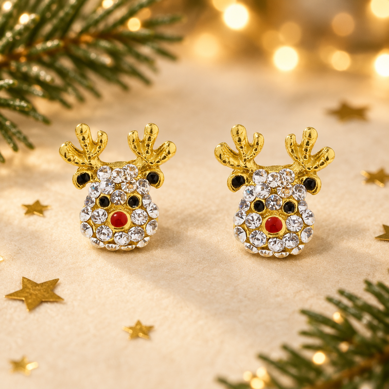 Pair of gold plated crystal Rudolph earrings laid face-up on a soft cream surface, surrounded by gold star confetti and evergreen sprigs with warm golden bokeh fairy lights in the background