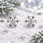 Pair of rhodium plated Cubic Zirconia snowflake earrings laid face-up in soft snow, surrounded by silver star confetti and frosted evergreen sprigs with cool bokeh fairy lights in the background