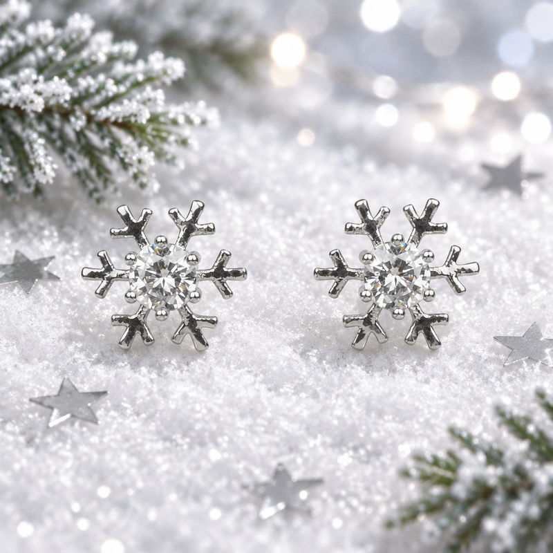 Pair of rhodium plated Cubic Zirconia snowflake earrings laid face-up in soft snow, surrounded by silver star confetti and frosted evergreen sprigs with cool bokeh fairy lights in the background
