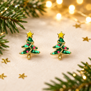 Pair of gold plated Christmas tree stud earrings laid face-up on a soft warm cream surface, surrounded by gold star confetti and evergreen sprigs with warm golden bokeh fairy lights in the background