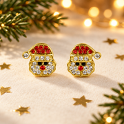 Pair of gold plated crystal Santa face earrings laid on a soft warm cream surface, surrounded by gold star confetti and evergreen sprigs with warm golden bokeh fairy lights in the background