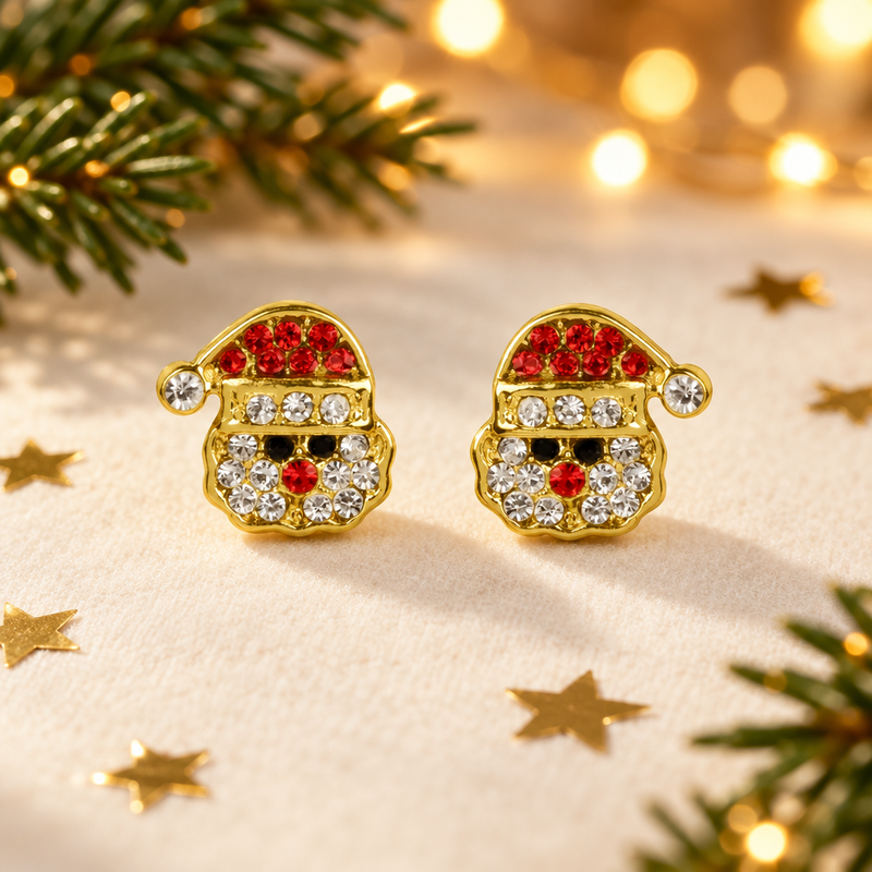 Pair of gold plated crystal Santa face earrings laid on a soft warm cream surface, surrounded by gold star confetti and evergreen sprigs with warm golden bokeh fairy lights in the background