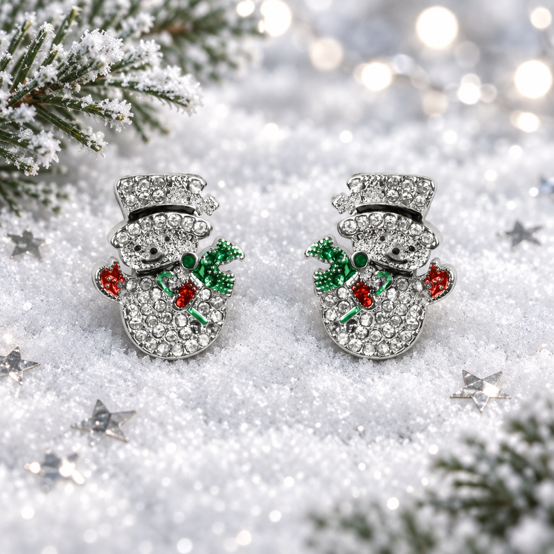 Pair of rhodium plated crystal snowman earrings lying flush in soft snow, surrounded by silver star confetti and frosted evergreen sprigs with cool bokeh fairy lights in the background