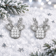 Pair of rhodium plated Cubic Zirconia reindeer head earrings lying flush in soft snow, surrounded by silver star confetti and frosted evergreen sprigs with cool bokeh fairy lights in the background