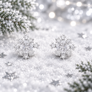 Pair of rhodium plated Cubic Zirconia snowflake earrings lying flush in soft snow, surrounded by silver star confetti and frosted evergreen sprigs with cool bokeh fairy lights in the background