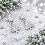 Pair of rhodium plated Cubic Zirconia star and Christmas tree drop earrings lying flush in soft snow, surrounded by silver star confetti and frosted evergreen sprigs with cool bokeh fairy lights in the background