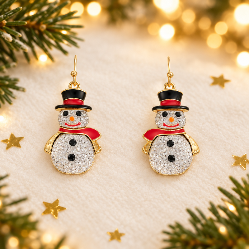 Pair of gold plated crystal snowman drop earrings laid on a soft warm white surface, surrounded by gold star confetti and evergreen sprigs with warm golden fairy lights in the background