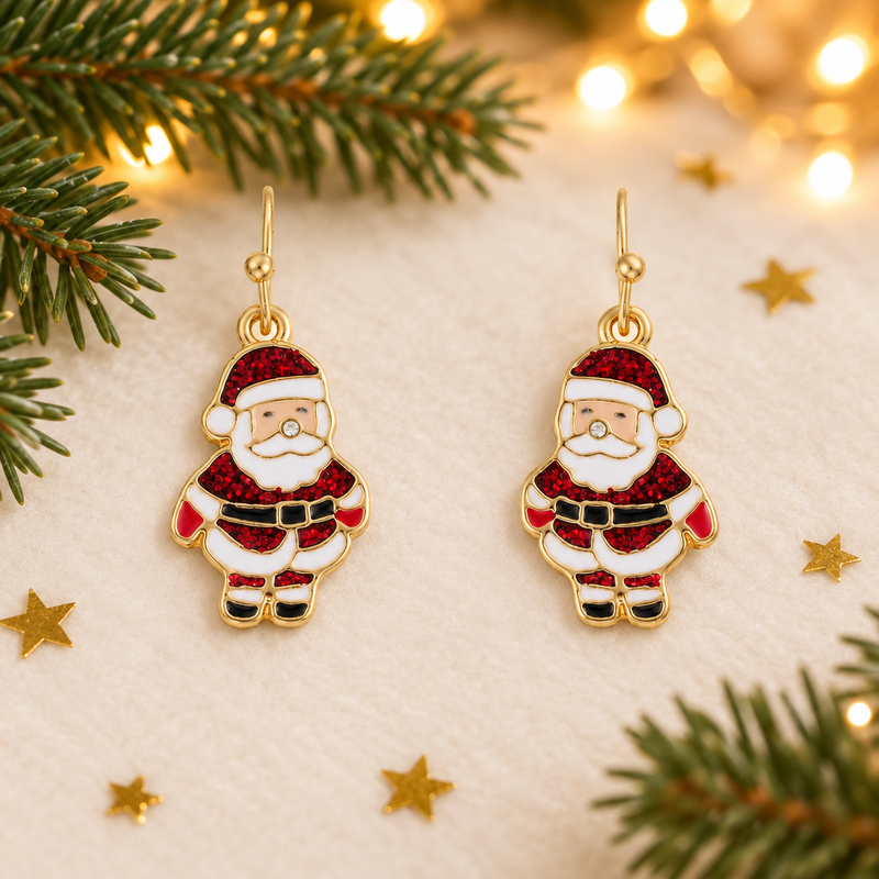 Pair of gold plated Santa drop earrings laid on a soft warm white surface, surrounded by gold star confetti and evergreen sprigs with warm golden fairy lights in the background