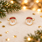 Pair of gold plated crystal-effect Santa stud earrings laid on a soft warm white surface, surrounded by gold star confetti and evergreen sprigs with warm golden fairy lights in the background