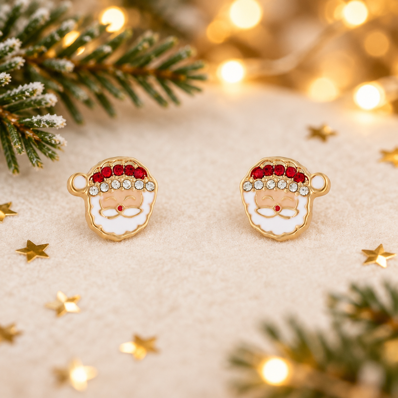Pair of gold plated crystal-effect Santa stud earrings laid on a soft warm white surface, surrounded by gold star confetti and evergreen sprigs with warm golden fairy lights in the background