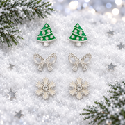 Trio of silver plated tree, bow and snowflake stud earrings laid face-up in soft snow, surrounded by silver star confetti and frosted evergreen sprigs with cool bokeh fairy lights in the background