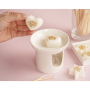 Heart-shaped ivory wax melt with gold star confetti being placed into white wax melt burner, The Little Things Mum collection on pink background