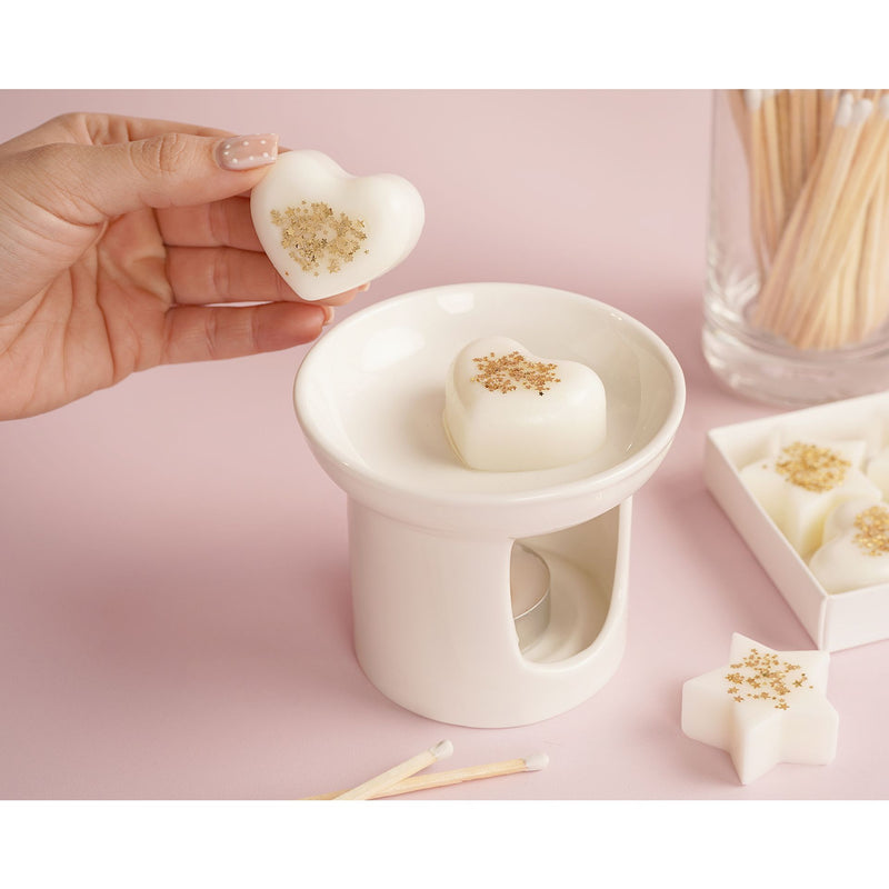 Heart-shaped ivory wax melt with gold star confetti being placed into white wax melt burner, The Little Things Mum collection on pink background