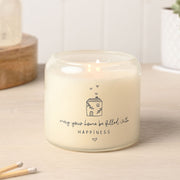 Moments Happy Home double wick candle in frosted glass jar with illustrated cottage and floating hearts design, sentiment reads may your home be filled with Happiness, lit on wooden surface