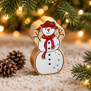 Wooden snowman toy with robin detail beneath Christmas tree branches beside small pinecones and warm fairy lights.