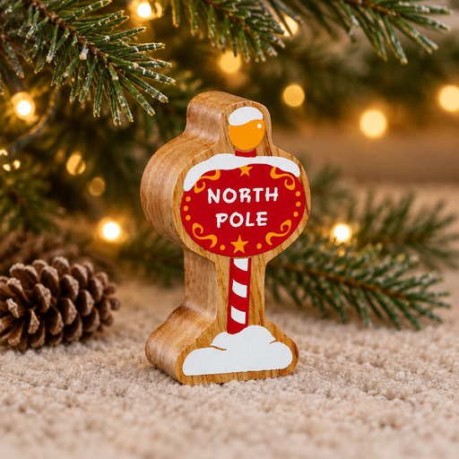 Wooden North Pole sign toy beneath Christmas tree branches beside a pinecone and warm fairy lights