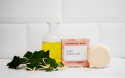 Laneside Soap Co Rose Geranium Shampoo Bar 60g with its box styled on a bathroom shelf with green foliage and a glass bottle of oil