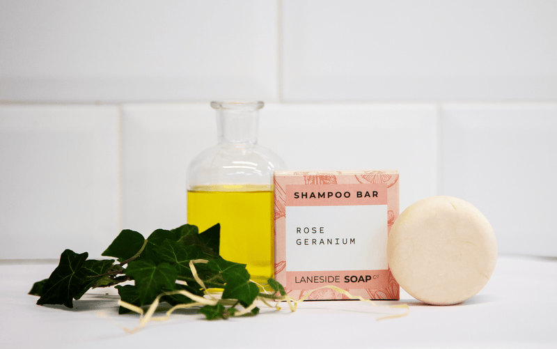Laneside Soap Co Rose Geranium Shampoo Bar 60g with its box styled on a bathroom shelf with green foliage and a glass bottle of oil
