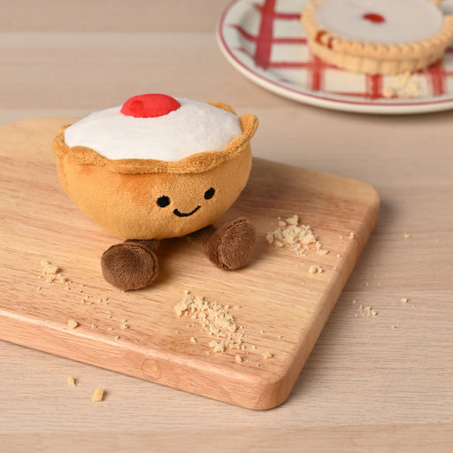 A soft plush Bakewell tart toy with a golden pastry case, white icing top, fluffy red cherry and smiling face, sitting on a wooden chopping board with crumbs scattered around and another Bakewell tart on a plate in the background