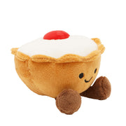Side angled view of a soft plush Bakewell tart toy with a golden pastry case with crimped edge detail, smooth white icing top, fluffy red cherry and fluffy brown feet, on a white background