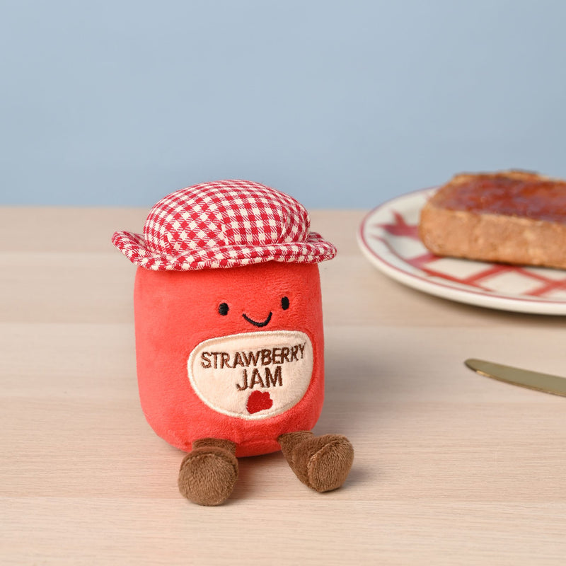 A soft red plush strawberry jam jar toy with a stitched Strawberry Jam label, smiling face and red and white gingham hat, sitting on a wooden table with a plate of toast spread with jam and a knife in the background against a soft blue wall