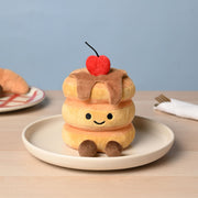 A soft plush pancake stack toy with three golden pancakes, syrup drizzle, a fluffy red cherry on top and small fluffy brown feet, sitting on a cream plate on a wooden table with a soft blue background