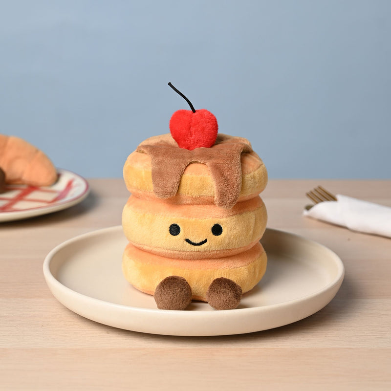 A soft plush pancake stack toy with three golden pancakes, syrup drizzle, a fluffy red cherry on top and small fluffy brown feet, sitting on a cream plate on a wooden table with a soft blue background