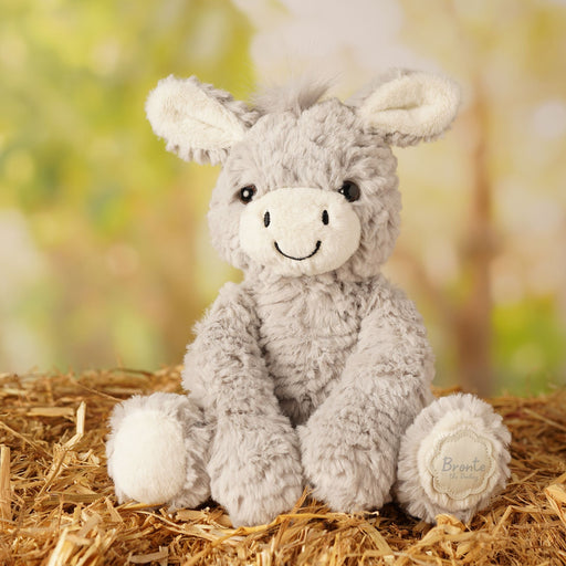 Bronte plush sitting donkey nestled in a bed of straw, lifestyle shot  with blurred background of trees
