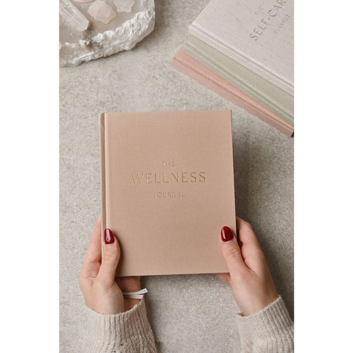 Serenity Wellness Journal held in two hands showing linen-effect blush hardcover with gold foil embossed title, lifestyle shot on stone surface