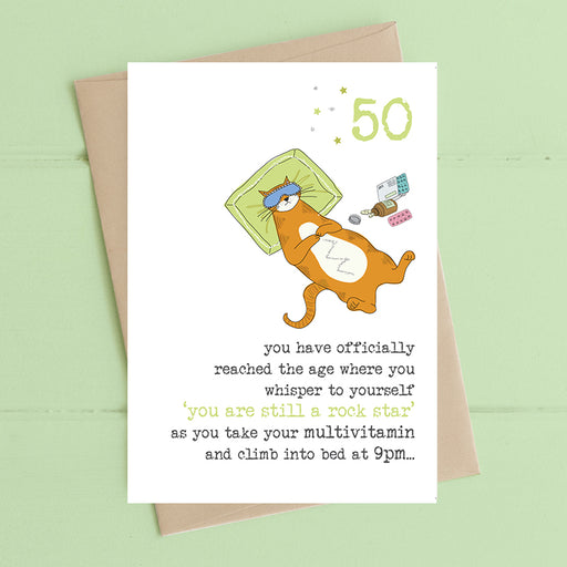Funny 50th birthday card with an illustrated cat sleeping in bed wearing an eye mask, surrounded by multivitamins, with the message "you are still a rock star as you take your multivitamin and climb into bed at 9pm"