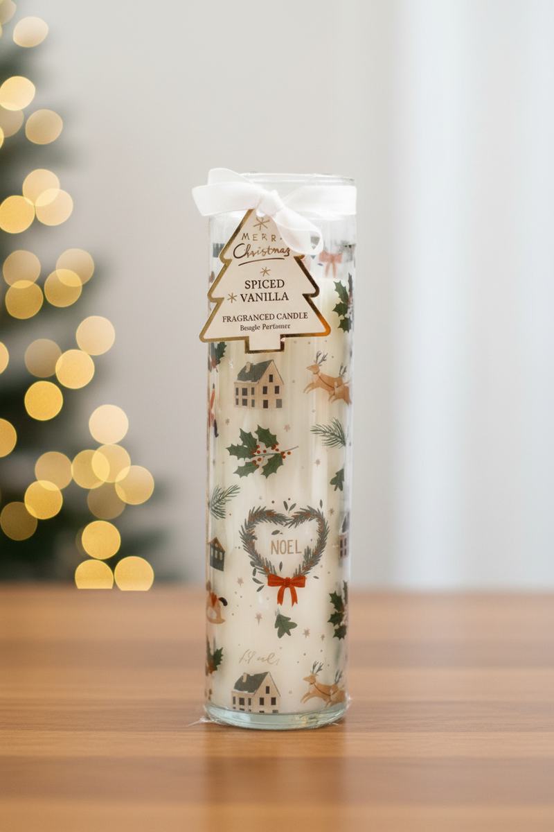 A tall glass tube candle with a Christmas scene featuring reindeer, holly, houses and a heart-shaped Noel wreath with a red bow, with a white ribbon and gold and cream Christmas tree gift tag, standing upright on a wooden surface in a warm festive setting with soft golden bokeh fairy lights in the background