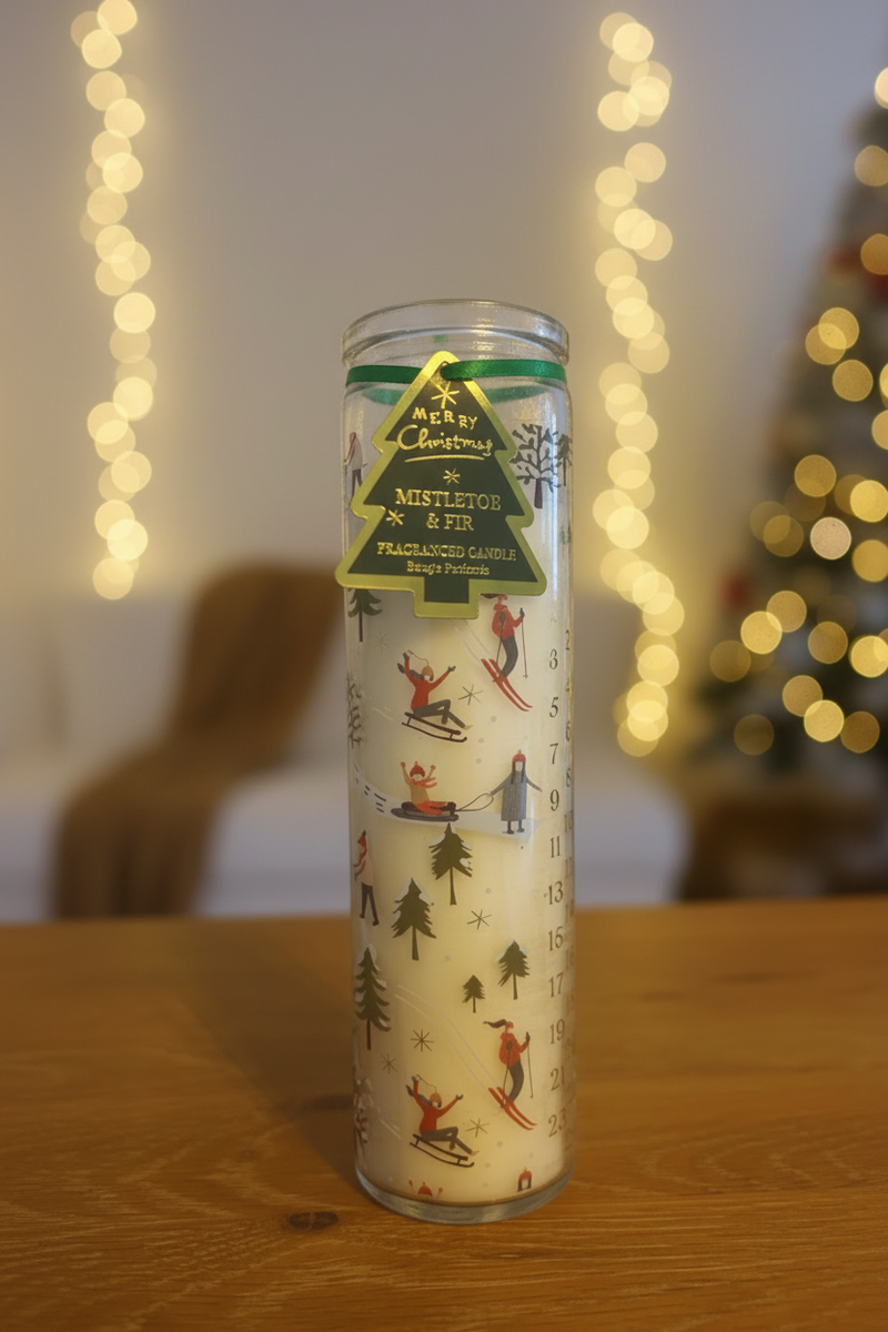 A tall glass tube candle with a winter scene featuring figures in the snow amongst green fir trees, with a bright green ribbon and green and gold Christmas tree gift tag, standing upright on a wooden surface in a warm festive setting with soft golden bokeh fairy lights in the background