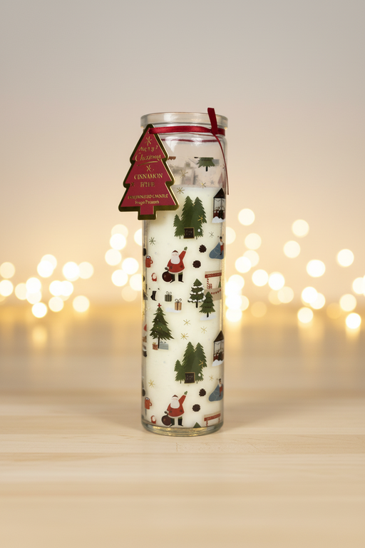 A tall glass tube candle with a Christmas village scene featuring Santa Claus, Christmas trees, snowflakes and gift boxes, with a dark red ribbon and Christmas tree gift tag, standing upright on a wooden surface in a warm festive setting with soft golden bokeh fairy lights in the background